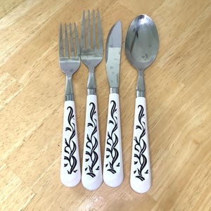 May include: A set of four pieces of flatware including a small fork, a large fork, a knife, and a spoon. Each piece features a polished silver-tone metal head and a white handle decorated with a black vine and leaf pattern running down the center.