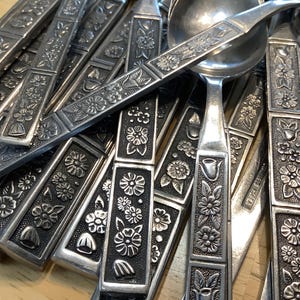 以下が含まれることがあります： ヴィンテージの銀食器のコレクションのクローズアップ。スプーンとフォークは、ハンドルに装飾的な花の模様があります。金属はくすんだ銀色で、花の細部がはっきりと見えます。