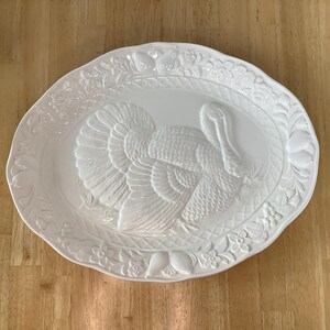 May include: An oval white ceramic serving platter featuring a raised, embossed turkey design in the center. The rim is decorated with a detailed relief pattern of fruits, leaves, and flowers, with a woven basket-weave texture surrounding the turkey.