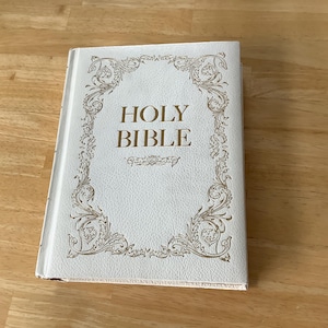 Puede incluir: Una Santa Biblia de tapa dura blanca con detalles dorados y las palabras "HOLY BIBLE" en letras doradas. El libro está sobre una superficie de madera.