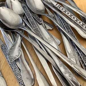 Può includere: Una collezione di cucchiai d'argento vintage con vari disegni del manico, tra cui motivi floreali e geometrici. I cucchiai sono disposti su una superficie di legno chiaro, mostrando le loro superfici riflettenti.