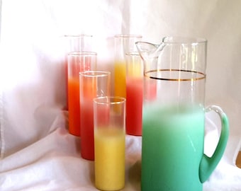 Pode incluir: Uma coleção de artigos de vidro vintage, incluindo copos altos e finos com gradientes de cores laranja, amarelo e vermelho, e um jarro grande com base verde e borda dourada. Os artigos de vidro estão dispostos sobre uma superfície branca.
