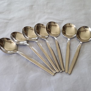 May include: Seven stainless steel soup spoons arranged on a white surface. Each spoon has a rounded bowl and a long, slender handle. The spoons reflect the light, creating a shiny appearance. Ideal for serving soups and stews.