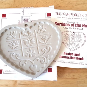 May include: A heart-shaped clay cookie mould with floral and butterfly designs, labelled "Gardens of the Heart." The mould is accompanied by a recipe and instruction book from The Pampered Chef.