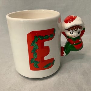 Könnte beinhalten: Weiße Keramiktasse mit einem roten Buchstaben "E", verziert mit grünem Stechpalmenlaub und Beeren. Der Griff ist eine Figur eines Kindes in einem roten und grünen Elfenoutfit und -hut. Eine festliche Tasse zum Thema Weihnachten.
