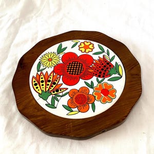 Puede incluir: Un salvamanteles decorativo con marco de madera y centro de baldosa cerámica. La baldosa presenta un diseño floral colorido con flores rojas, naranjas y amarillas sobre un fondo blanco. El marco de madera tiene un borde ondulado.