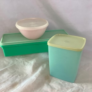 May include: A collection of vintage plastic food storage containers in various shapes and colors. Includes a pink bowl with a white lid, a green rectangular container, and a teal square container with a yellow lid. The containers are arranged on a white surface.