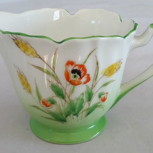 May include: A white ceramic creamer with a green rim and base. It features a floral design with red poppies, yellow wheat, and green leaves.