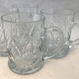 May include: Four clear glass mugs with a detailed, cut-glass pattern. Each mug has a rounded handle and a sturdy base. The light reflects off the facets, creating a sparkling effect. The mugs are arranged closely together.