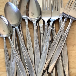 Puede incluir: Una colección de cubiertos antiguos, que incluye cucharas, tenedores y cuchillos, dispuestos sobre una superficie de madera clara. La cubertería tiene un acabado metálico brillante con un diseño grabado en los mangos. La disposición sugiere un juego de comedor.