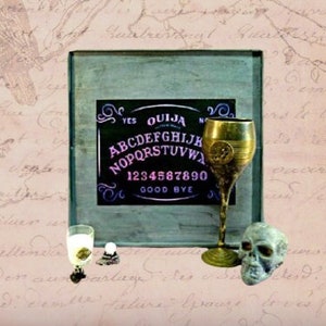 May include: A vintage Ouija board game set with a wooden frame, a black planchette, and a gold-coloured goblet. The board features the letters of the alphabet, numbers 0-9, and the words "yes", "no", "goodbye", and "ouija".