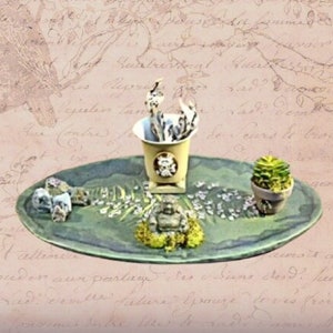 May include: A green ceramic tray with a white ceramic planter, a small potted succulent, and a small Buddha statue. The tray is decorated with dried flowers and small white stones.