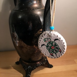 May include: A silver teapot with a decorative handle and a round, white leather ornament with a painted green turtle design hanging from it.