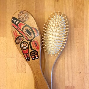 May include: Two bamboo hairbrushes. One brush features a Native American-inspired design in red, black, and white. The other brush has wooden bristles set in a white cushion. Both brushes have a natural wood handle.