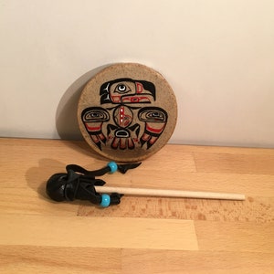 May include: A round, brown, wooden hand drum with a painted design of a Native American-style bird with red, black, and white details. A black leather drum stick with blue beads is included.