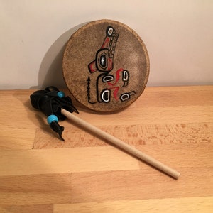 Op de afbeelding: Een ronde, bruine leren trommel met een geschilderd ontwerp van een rood en zwart wezen. Een zwarte en blauwe stok met een blauwe kraal ligt op een houten oppervlak.