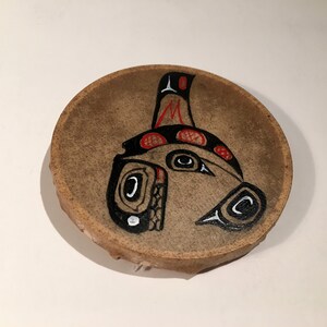 May include: A round, brown leather drum with a painted design of a black and white creature with red accents. The creature has large eyes and a long, pointed beak.