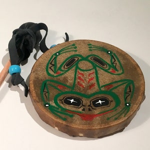 May include: A small, hand-crafted drum with a green frog design painted on the front. The drum is made of leather and has a wooden stick with a leather handle.