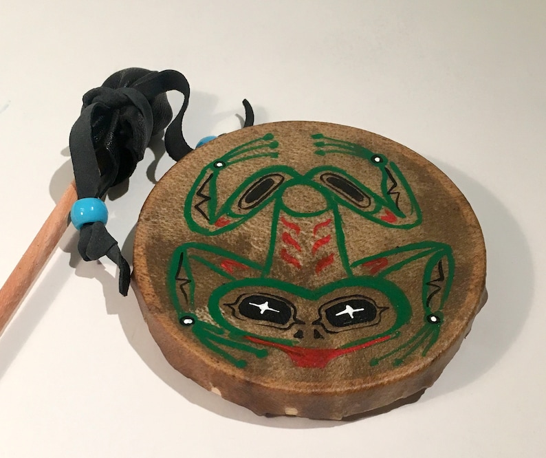 May include: A small, hand-crafted drum with a green frog design painted on the front. The drum is made of leather and has a wooden stick with a leather handle.
