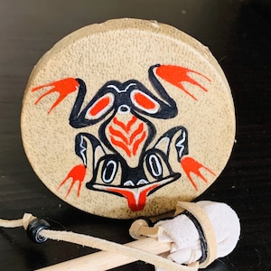 May include: A small hand drum with a painted design of a frog in black, red, and white. The drum is made of wood and has a leather head. It comes with a wooden stick with a white padded end.