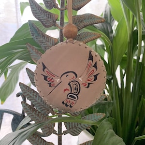 May include: A round, beige leather ornament with a painted hummingbird design. The hummingbird is red, black, and white with a stylized design. The ornament is hanging from a brown cord with a wooden bead.