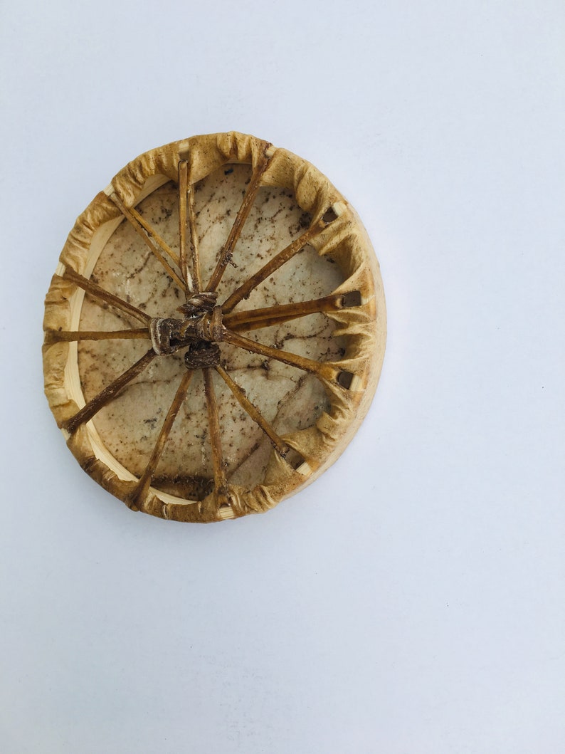 May include: A round, tan-colored Native American hand drum with a leather covering and a woven design. The drum is made of wood and has a natural, rustic look.