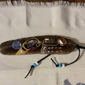 Puede incluir: Una pluma marrón con un diseño pintado de un pájaro en negro, blanco, dorado y rojo. La pluma está atada con un cordón negro y tiene cuentas azules y blancas.