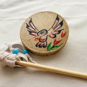 May include: A wooden hand drum with a painted design of a red, white and black bird with green leaves and red flowers. The drum is attached to a wooden stick with a white leather strap and blue beads.