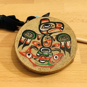 Könnte beinhalten: Eine kleine, runde, hölzerne Handtrommel mit einem bemalten Design eines stilisierten Vogels mit roten, grünen, schwarzen und weißen Details. Die Trommel wird von einem Holzstock begleitet.