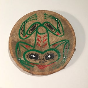 May include: A round, brown leather drum with a painted green frog design. The frog has black eyes, red markings, and white accents.