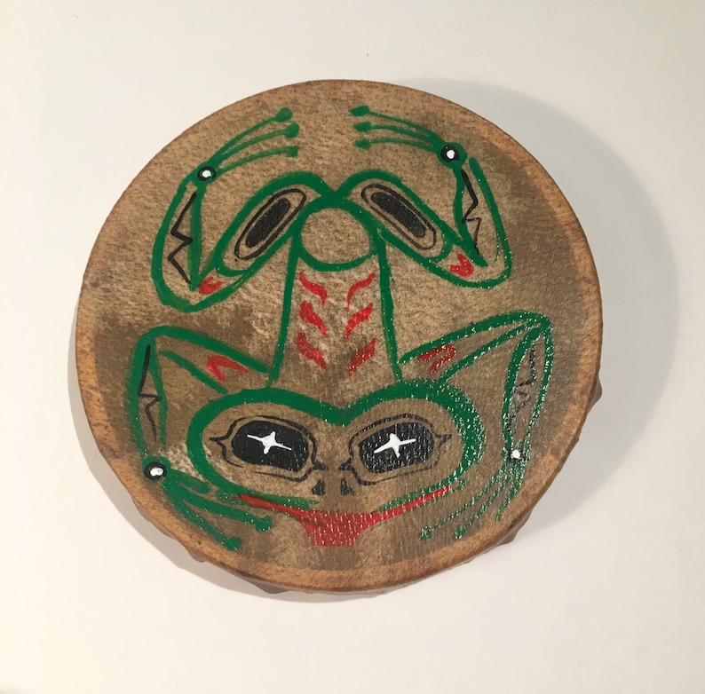 May include: A round, brown leather drum with a painted green frog design. The frog has black eyes, red markings, and white accents.