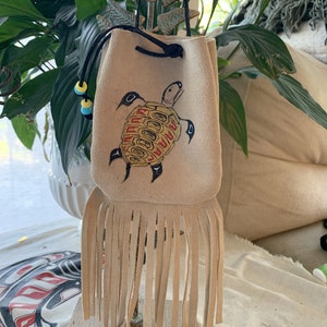 May include: A beige leather pouch with a fringe bottom and a drawstring closure. The pouch is decorated with an embroidered image of a turtle with yellow and red accents.