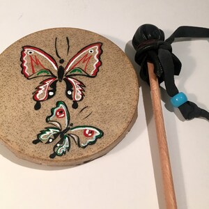 Könnte beinhalten: Eine kleine, runde, braune Ledertrommel mit zwei bemalten Schmetterlingen. Der obere Schmetterling ist rot, weiß und grün. Der untere Schmetterling ist grün, weiß und schwarz. Die Trommel wird mit einem Holzstock mit einem schwarzen Lederhammer am Ende geliefert.