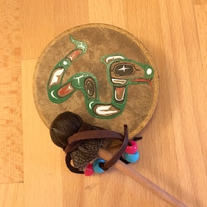 Könnte beinhalten: Eine kleine, runde, braune Ledertrommel mit einem bemalten grünen Schlangenmotiv. Die Trommel wird mit einem braunen Lederhammer mit einem Holzstiel und blauen und rosa Perlen geliefert.