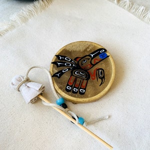 May include: A small, round, hand-painted drum with a design of a bird with outstretched wings. The drum is made of wood and has a leather top. The bird is painted in black, red, and blue. The drum is on a white fabric surface.