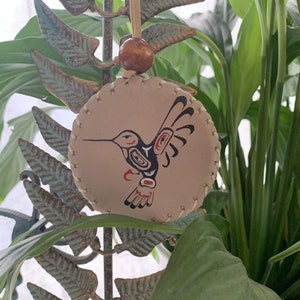 May include: A beige leather ornament featuring a hand-painted hummingbird design. The hummingbird is black, red and white with a stylised design. The ornament is hanging from a brown cord.