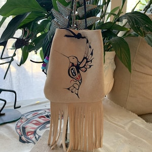 May include: A beige leather pouch with a fringe bottom and a hummingbird design. The pouch is hanging from a black cord and has a drawstring closure.