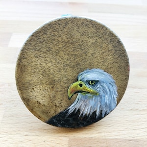 Könnte beinhalten: Ein handbemalter Holz-Untersetzer mit einer Nahaufnahme des Kopfes eines Weißkopfseeadlers mit gelbem Schnabel und weißen Federn. Das Auge des Adlers ist leuchtend gelb.