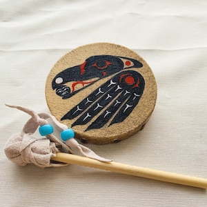 Könnte beinhalten: Eine kleine, runde, hölzerne Handtrommel mit einem schwarz-roten bemalten Design eines Vogels mit ausgebreiteten Flügeln. Die Trommel wird mit einem kleinen, hölzernen Schlägel mit einer braunen Lederummantelung und zwei türkisfarbenen Perlen geliefert.