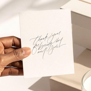 May include: A lit candle in a clear glass, a gift box tied with a ribbon, and a card with the text "Thank you for being by our side" are arranged on a white surface. The items are lit by natural light.