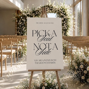 May include: A white rectangular wedding sign on a wooden easel featuring black text that reads: THE SMITHS - AUGUST 20, 2026, PICK A SEAT NOT A SIDE, YOU ARE LOVED BY BOTH THE GROOM AND BRIDE. The sign is displayed in a bright, open indoor ceremony space.