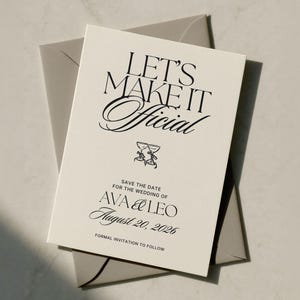 Może przedstawiać: Kremowa, prostokątna karta typu save the date z czarnym napisem LET'S MAKE IT Official, małą ilustracją dwóch kieliszków do szampana, imionami AVA & LEO oraz datą 20 sierpnia 2026 roku, umieszczona na szarej kopercie.