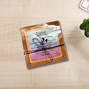 May include: A teacher's gift set, comprising a card with text and a decorative bow. The card features a colourful design with the word "Teacher" at the top. The text describes the qualities of a teacher. The set is packaged in a brown box.