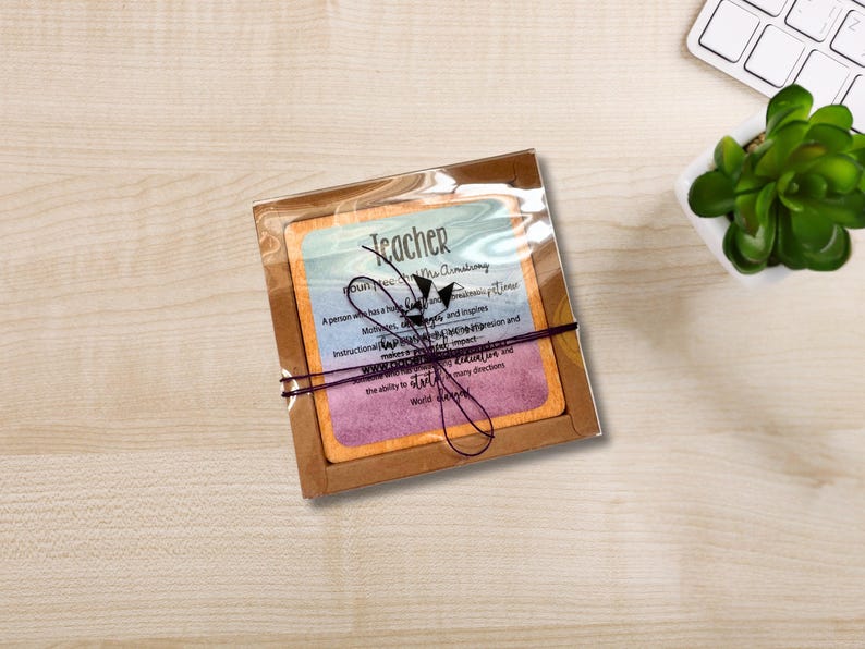 May include: A teacher's gift set, comprising a card with text and a decorative bow. The card features a colourful design with the word "Teacher" at the top. The text describes the qualities of a teacher. The set is packaged in a brown box.
