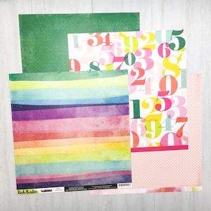 May include: A set of three patterned scrapbook papers. The first paper is green with a subtle pattern. The second paper has a rainbow stripe pattern. The third paper has a pink background with white polka dots and colorful numbers.