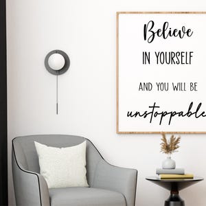 K&ouml;nnte beinhalten: Ein gerahmter Druck mit den Worten "Believe in Yourself and You Will Be Unstoppable" in schwarzer Schrift. Der Druck h&auml;ngt &uuml;ber einem grauen Sessel mit einem wei&szlig;en Kissen. Ein kleiner Beistelltisch enth&auml;lt B&uuml;cher und eine Vase mit getrockneten Blumen.