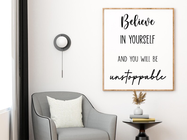 K&ouml;nnte beinhalten: Ein gerahmter Druck mit den Worten "Believe in Yourself and You Will Be Unstoppable" in schwarzer Schrift. Der Druck h&auml;ngt &uuml;ber einem grauen Sessel mit einem wei&szlig;en Kissen. Ein kleiner Beistelltisch enth&auml;lt B&uuml;cher und eine Vase mit getrockneten Blumen.