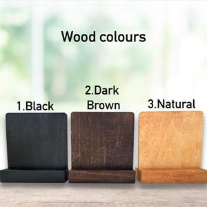 May include: Three wooden phone stands in black, dark brown, and natural colours. The stands are labelled "Wood colours" and numbered 1, 2, and 3. The background is a blurred view of a window.