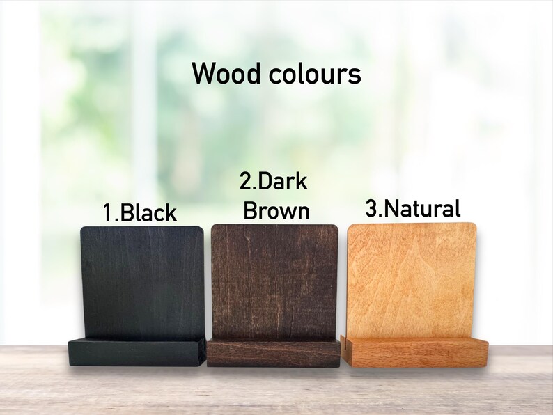 May include: Three wooden phone stands in black, dark brown, and natural colours. The stands are labelled "Wood colours" and numbered 1, 2, and 3. The background is a blurred view of a window.