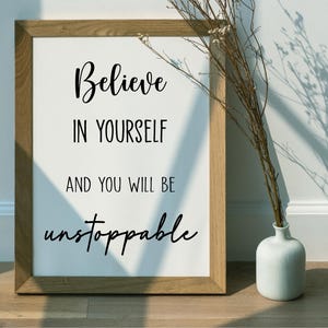 K&ouml;nnte beinhalten: Ein gerahmter Druck mit den Worten "Believe in Yourself and You Will Be Unstoppable" in schwarzer Schrift. Der Rahmen ist aus hellem Holz. Rechts vom Rahmen steht eine kleine wei&szlig;e Vase mit getrockneten Zweigen.